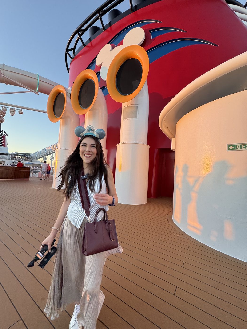 Dani Godinez con orejas de Mickey caminando por la cubierta de un barco de Disney Cruise Line, junto a la chimenea.