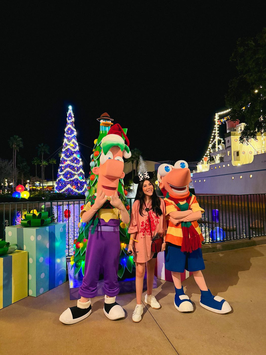 Dani Godinez con Phineas y Ferb, con ropa navideña, junto al árbol de Navidad en Jollywood Nights.